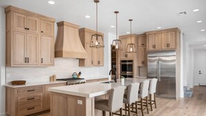 A modern kitchen with wooden cabinets, stainless steel appliances, and a central island with bar stools. - Desert Gem 7 Bedroom Home (St. George)