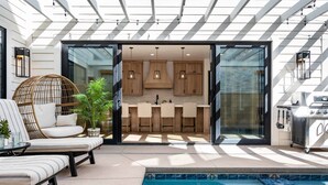 A modern outdoor kitchen with a glass door leading to a dining area, a pool, and lounge seating. - Desert Gem 7 Bedroom Home (St. George)