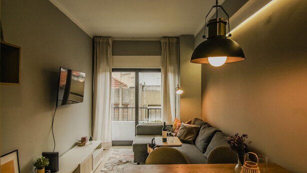 Ett modernt vardagsrum med en soffa, ett matbord i trä, en tv som är monterad på väggen och ett stort fönster med gardiner.