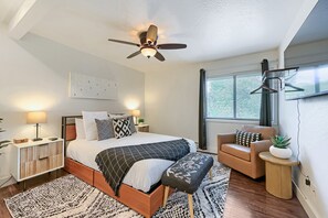 Ein modernes Schlafzimmer mit Bett, Nachttischen, Deckenventilator, Fenster mit Jalousien, einem Sessel, einem kleinen Tisch mit Pflanze und einem gemusterten Teppich.