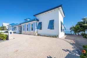 Ein modernes, weißes Gebäude mit blauen Fensterläden, einer gepflasterten Auffahrt und Blick auf den Strand.