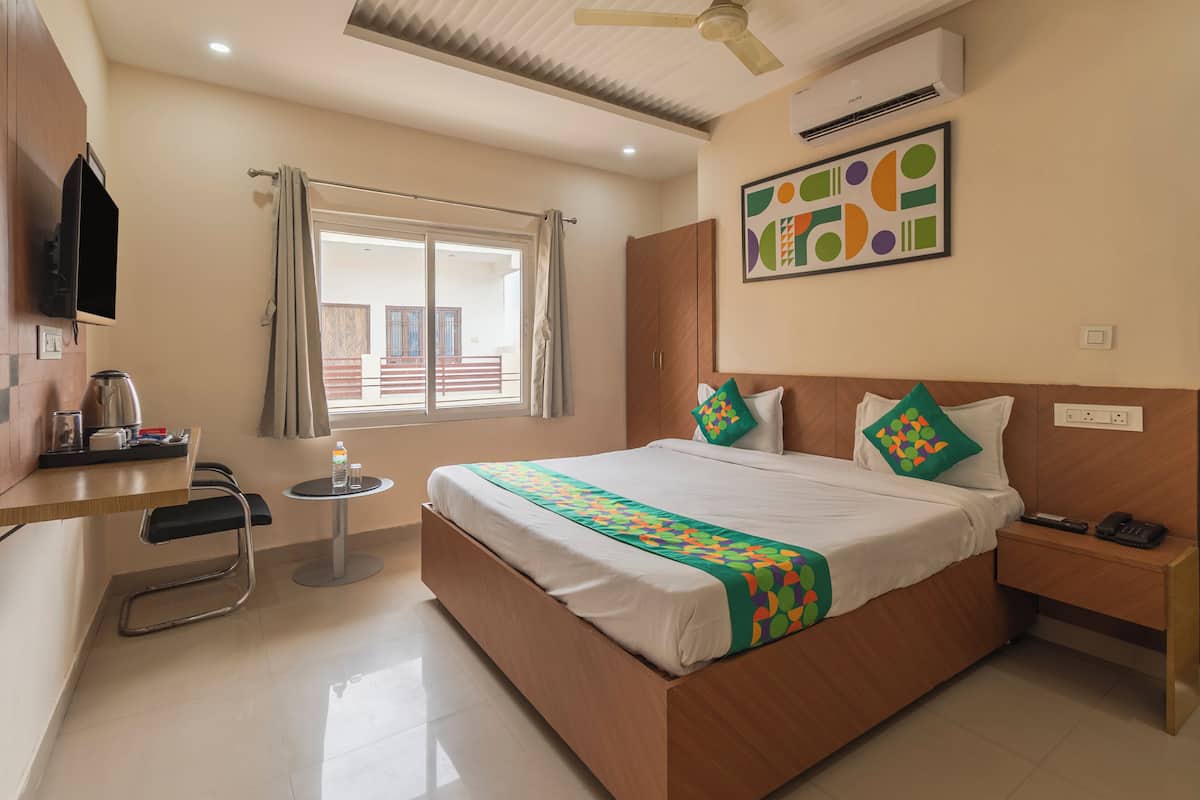 Habitación de hotel con cama, mesita de noche, una pequeña mesa redonda con una silla, televisor montado en la pared y una ventana con cortinas.