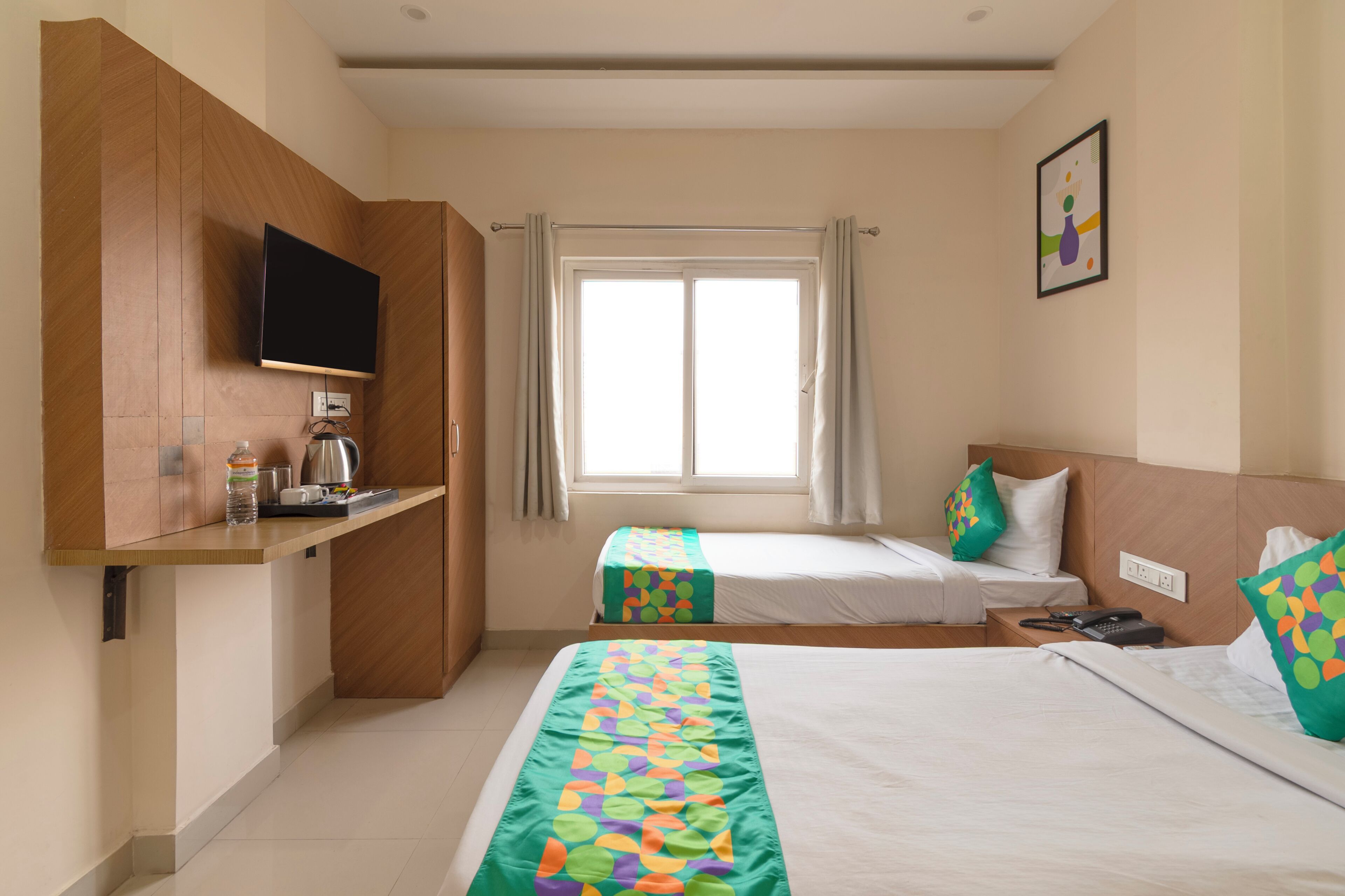 A hotel room with two beds, a TV mounted on the wall, a window with curtains, and a small kitchenette area.