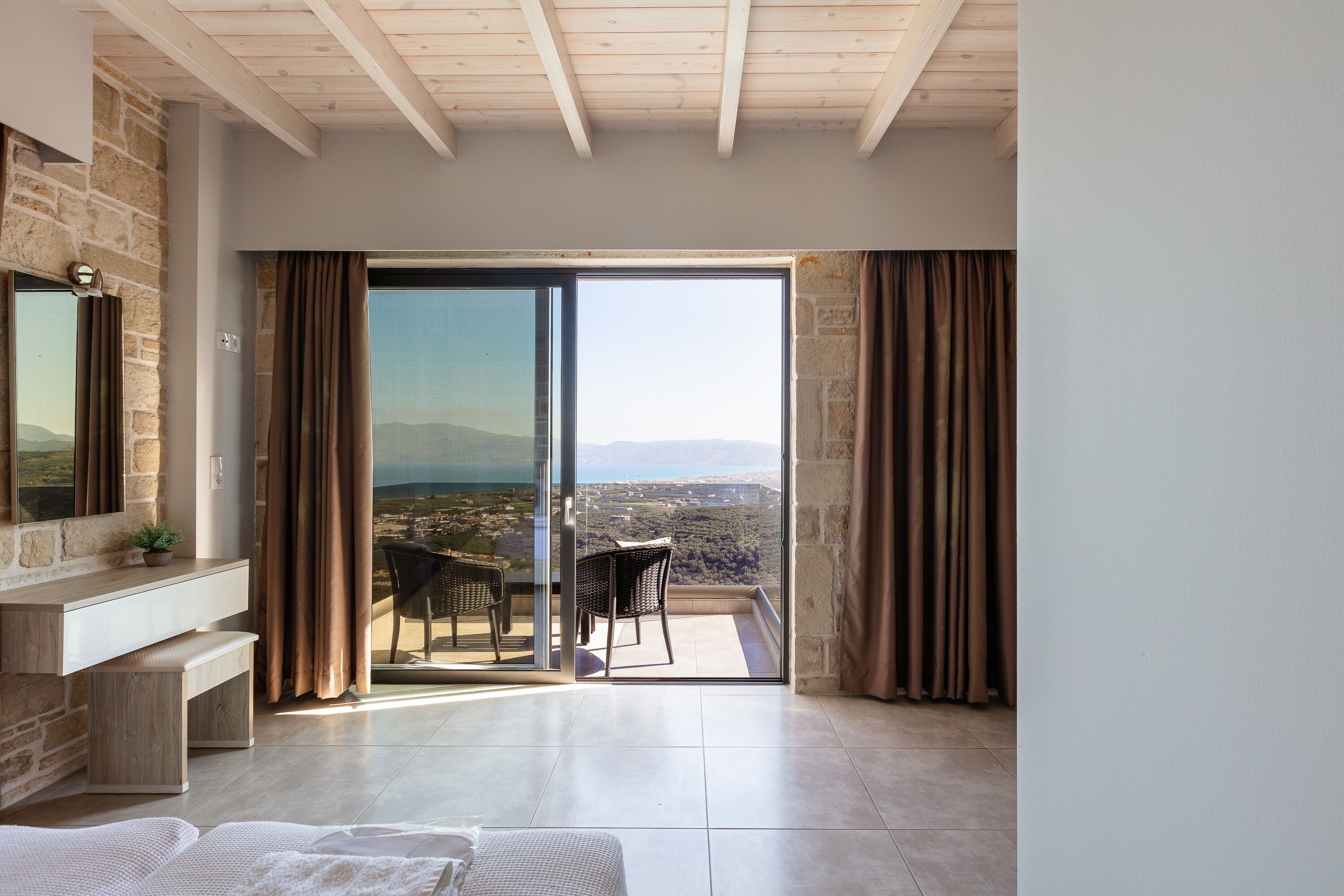 A spacious room with a balcony, a stone wall, a wooden bench, a bed, and a view of the landscape.