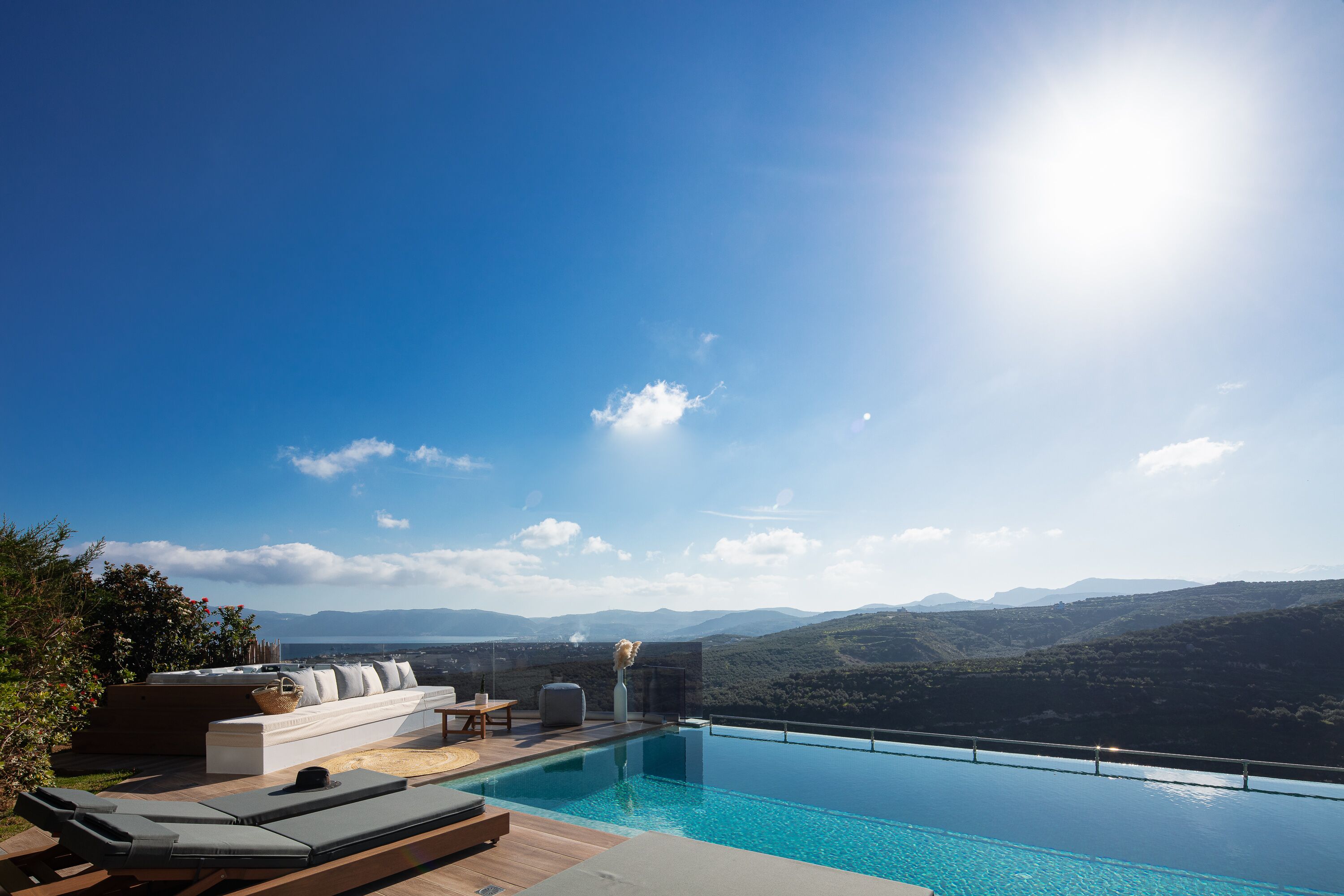 An infinity pool with a clear view of the mountains, a sun lounger, and a seating area with cushions and a vase.