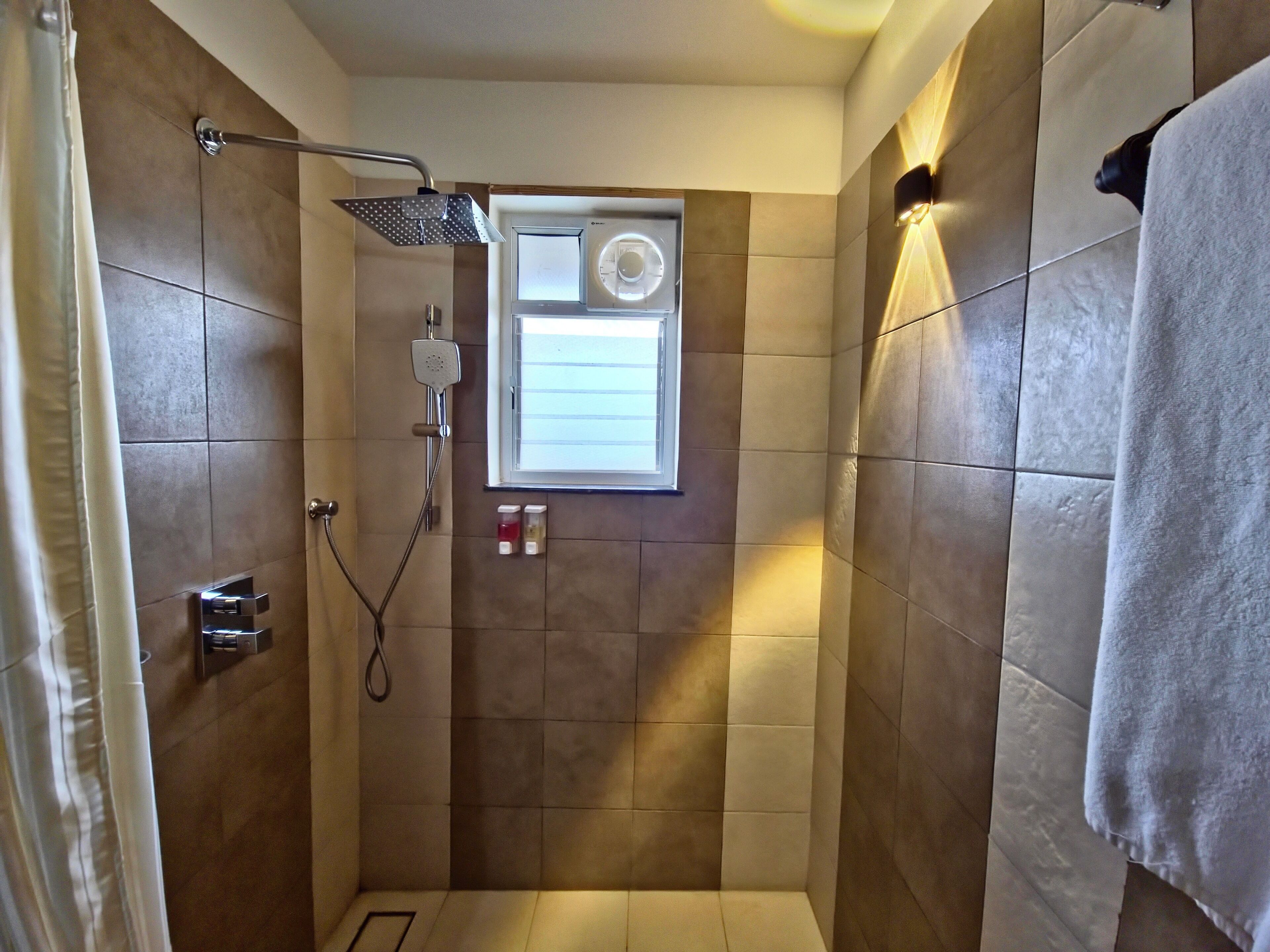 Une petite salle de douche carrelée, dotée d’une fenêtre, d’un porte-serviettes et d’un pommeau de douche.
