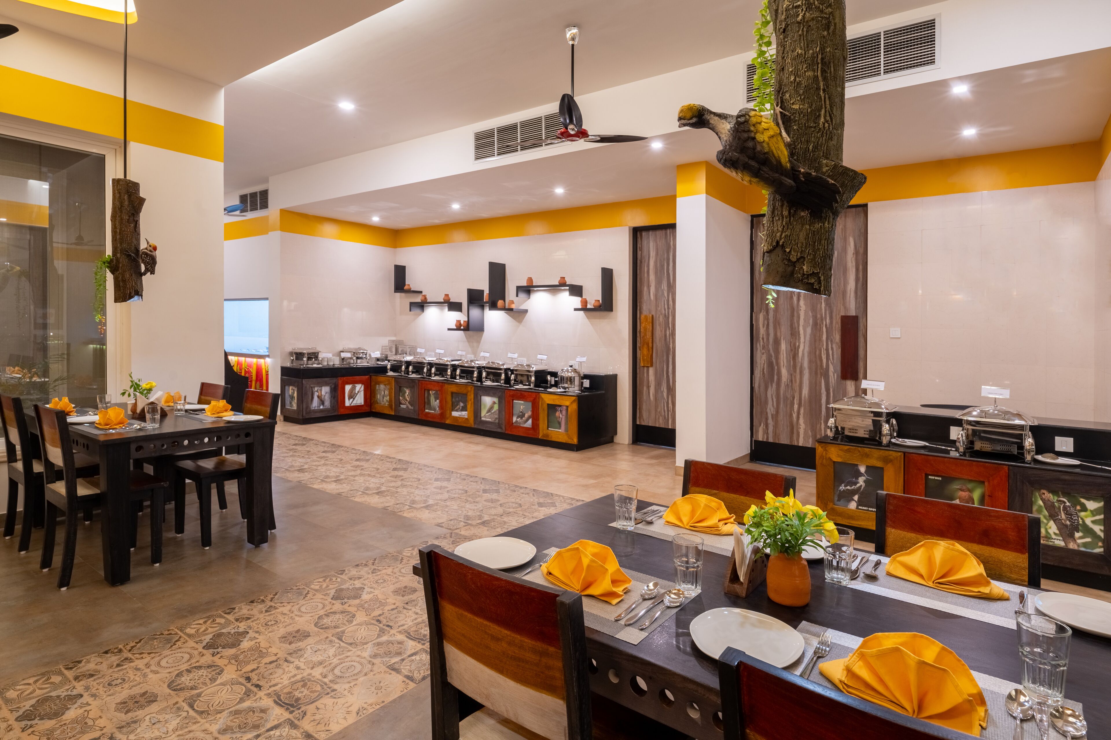 A dining area with tables set for four, a kitchen in the background, and a decorative tree trunk.
