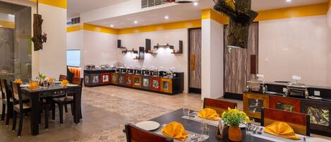 A dining area with tables set for four, a kitchen in the background, and a decorative tree trunk.