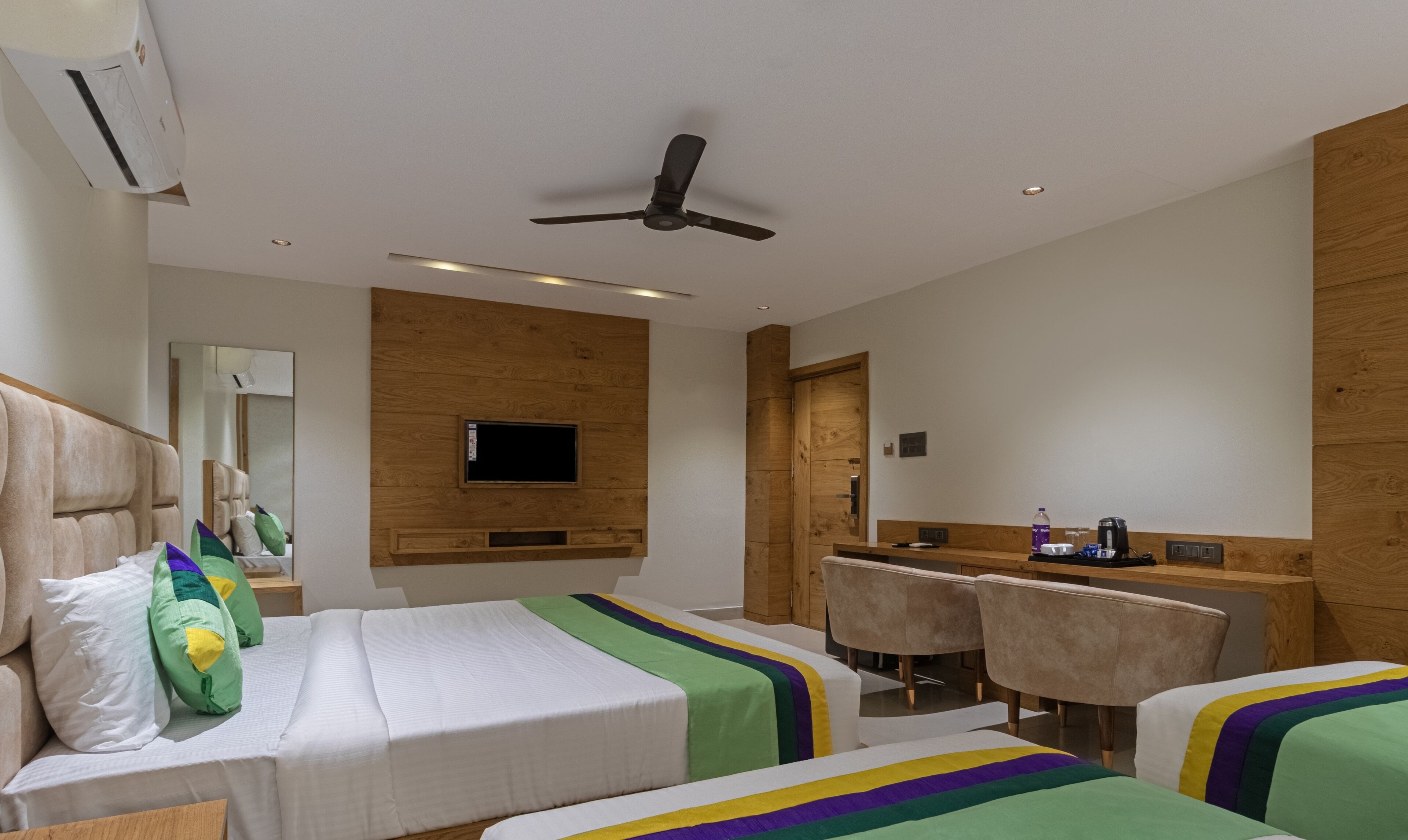 A hotel room with two beds, a desk, chairs, a television, and a ceiling fan.