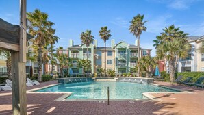 A pool area with lounge chairs and palm trees, surrounded by multi-story buildings with balconies. - Beautiful Ocean & Beach Front Condo~Sleeps 5 - Amazing Views! (Galveston)