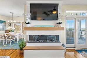 A living room with a TV, a fireplace, a dining area, and a view of the beach.