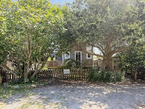 A rustic wooden cabin with a gravel driveway and a wooden fence. - Treehouse Charlie: Cute Cottage for Couples or Small Families (Ocracoke)