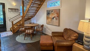 A living room with a wooden staircase, a round table with chairs, a brown leather sofa, a painting on the wall, and a lamp on a side table. - Treehouse Charlie: Cute Cottage for Couples or Small Families (Ocracoke)