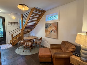 A living room with a wooden staircase, a round table with chairs, a brown leather sofa, a painting on the wall, and a lamp on a side table. - Treehouse Charlie: Cute Cottage for Couples or Small Families (Ocracoke)