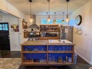 Eine Küche mit einer zentralen Kochinsel, Holzregalen, hängenden Pendelleuchten, einem Herd, einer Mikrowelle, einem Kühlschrank und einer Wanduhr.