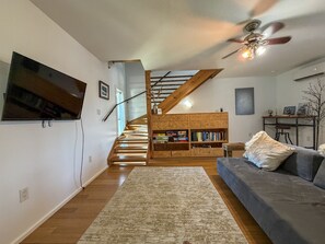 Ein Wohnzimmer mit einem Flachbildfernseher, einer Holztreppe, einem Bücherregal, einem blauen Sofa und einem Deckenventilator.