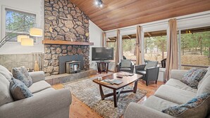 A cozy living room with a stone fireplace, comfortable seating, and large windows offering a view of the outdoors. - Forest Retreat with Access to The Cove (Sunriver)