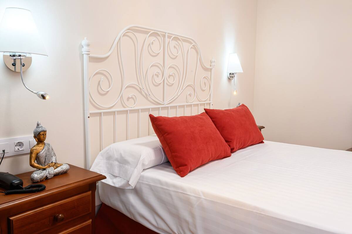 A neatly made bed with red pillows, a white headboard, and a bedside table with a lamp and a statue.