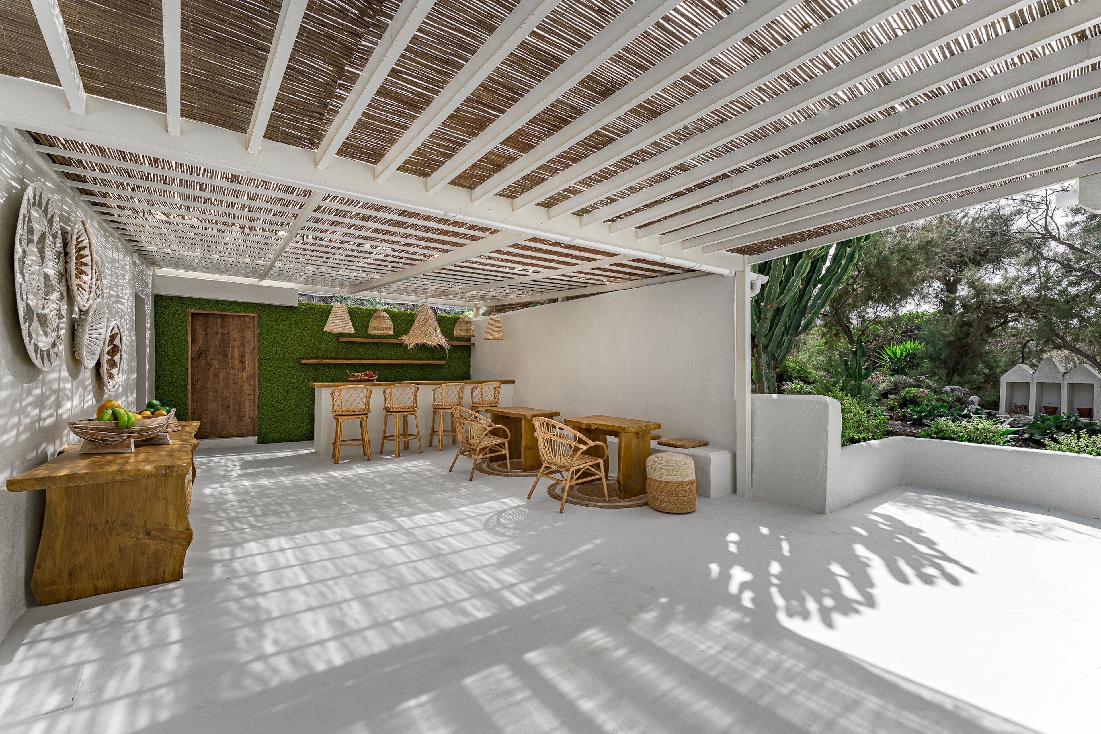 A spacious patio with a wooden table and chairs, a green wall, and a view of greenery outside.