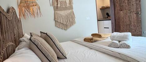 Una cama bien hecha con sábanas blancas y un cabecero de madera, un armario de madera y una lámpara de macramé fijada a la pared.
