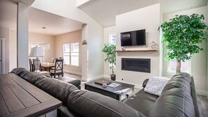 A modern living room with a sofa, coffee table, and fireplace. There is a television mounted above the fireplace and a dining area in the background.