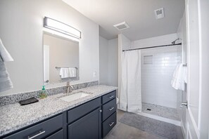 Un baño moderno con lavamanos de granito, muebles oscuros, un espejo grande y una ducha a ras de suelo con puerta de cristal.