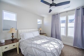 Un dormitorio con una cama, dos mesitas de noche, un ventilador de techo y ventanas con cortinas.