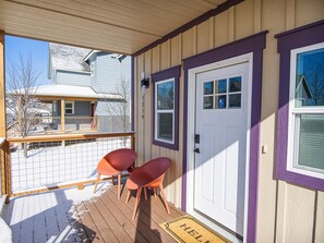 A covered porch with a wooden deck, two red chairs, and a white door with a blue sign.