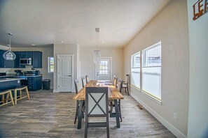 A spacious dining area with a long wooden table and chairs, a kitchen with dark blue cabinets, and large windows providing natural light.