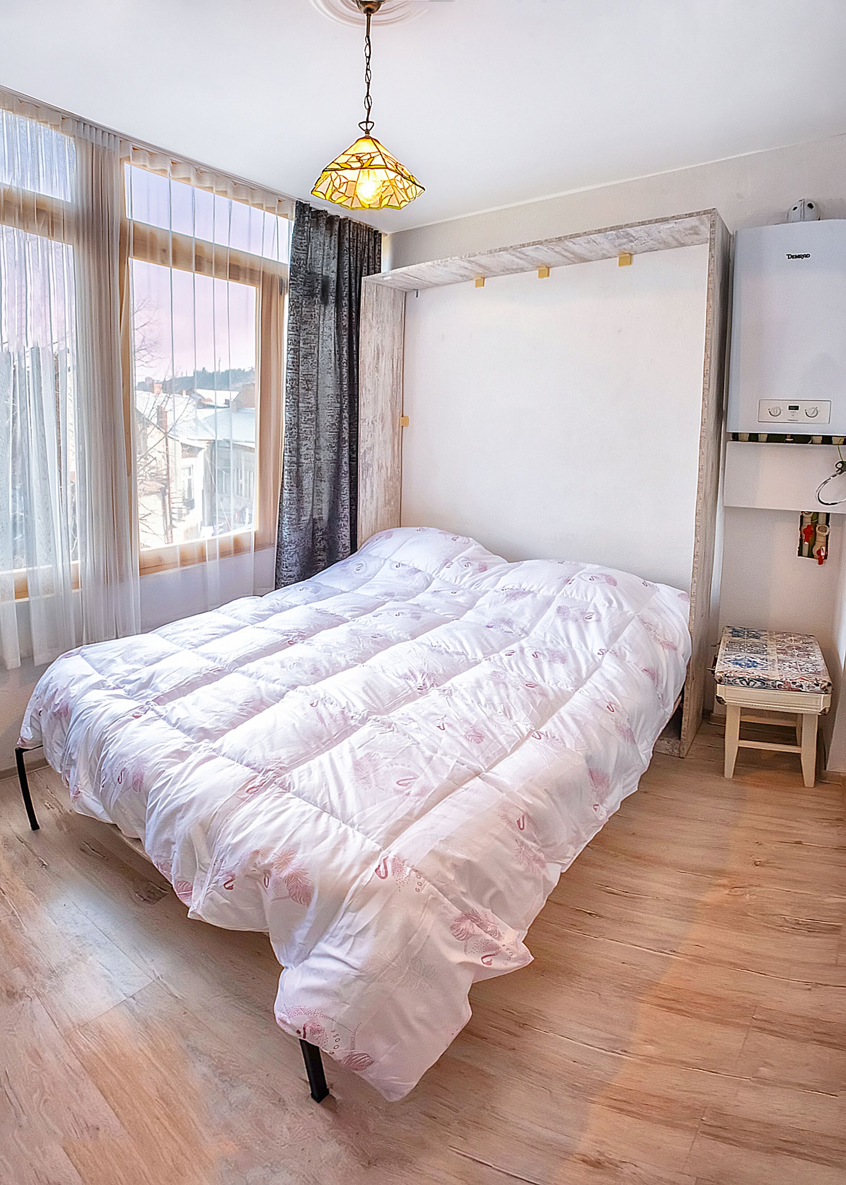 Une chambre à coucher avec un grand lit, une petite table, un luminaire au plafond et une fenêtre avec des rideaux.