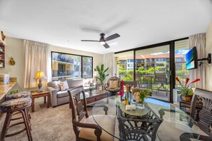 A spacious living area with a glass coffee table, comfortable seating, a ceiling fan, and a view of a residential building.