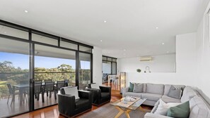 Un soggiorno moderno con divano, tavolino e poltrone, una zona pranzo con tavolo e sedie, e un'ampia porta scorrevole in vetro che dà su un balcone con vista sul verde.