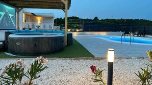Un espace piscine comprenant un jacuzzi, un coin salon couvert et un grand écran pour les divertissements.