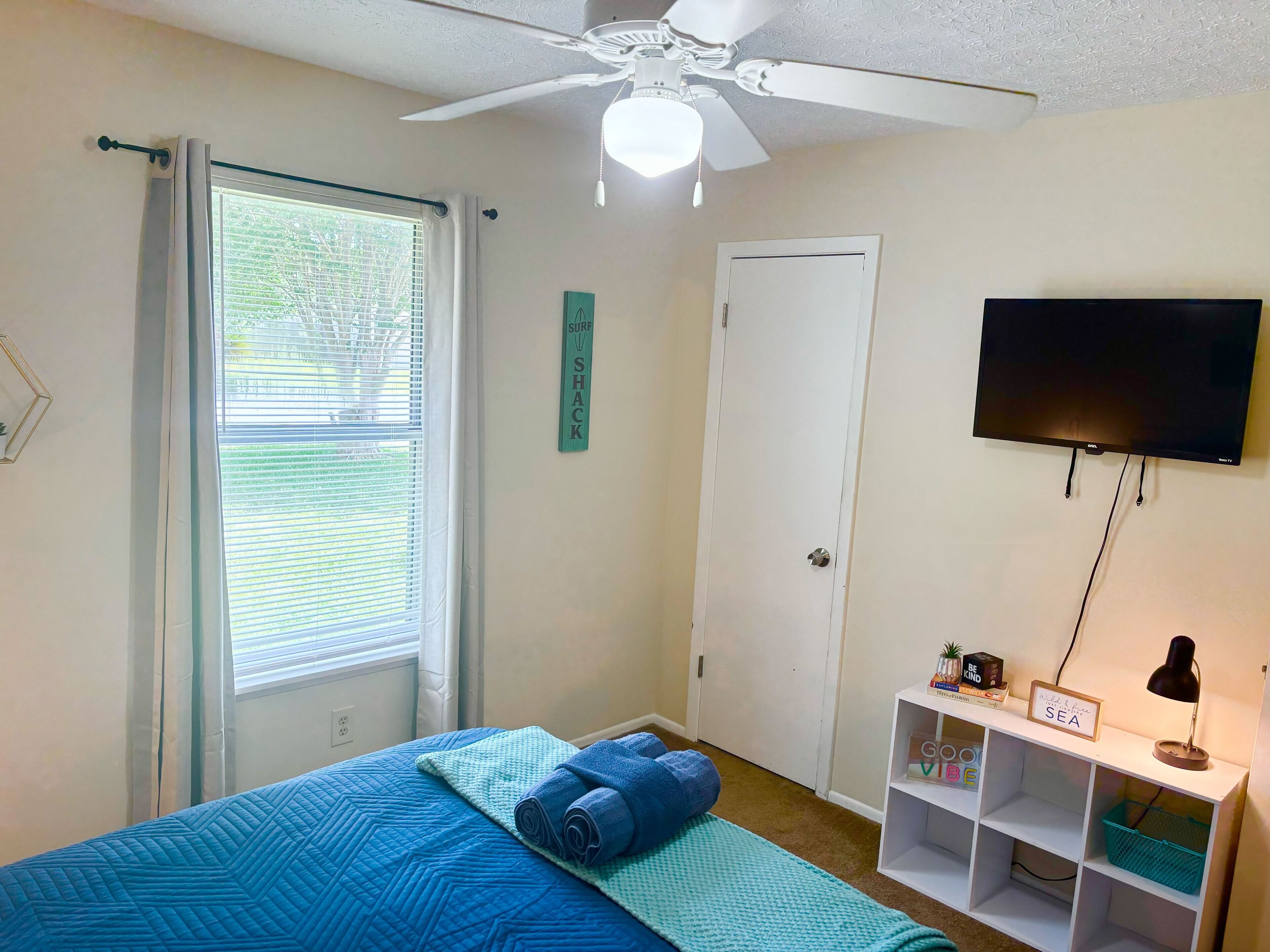 A bedroom with a bed, a ceiling fan, a TV, and a window with blinds.