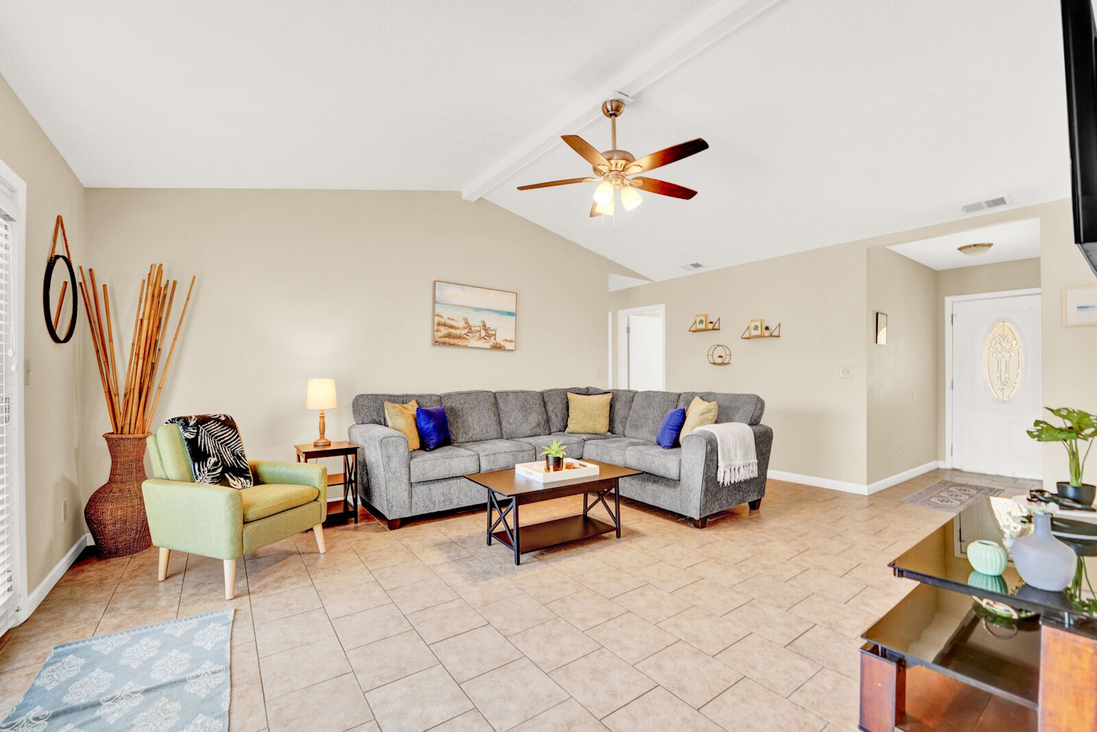 A spacious living room with a grey sectional sofa, a coffee table, and a ceiling fan. There is a door leading to another room and a window on the left side.