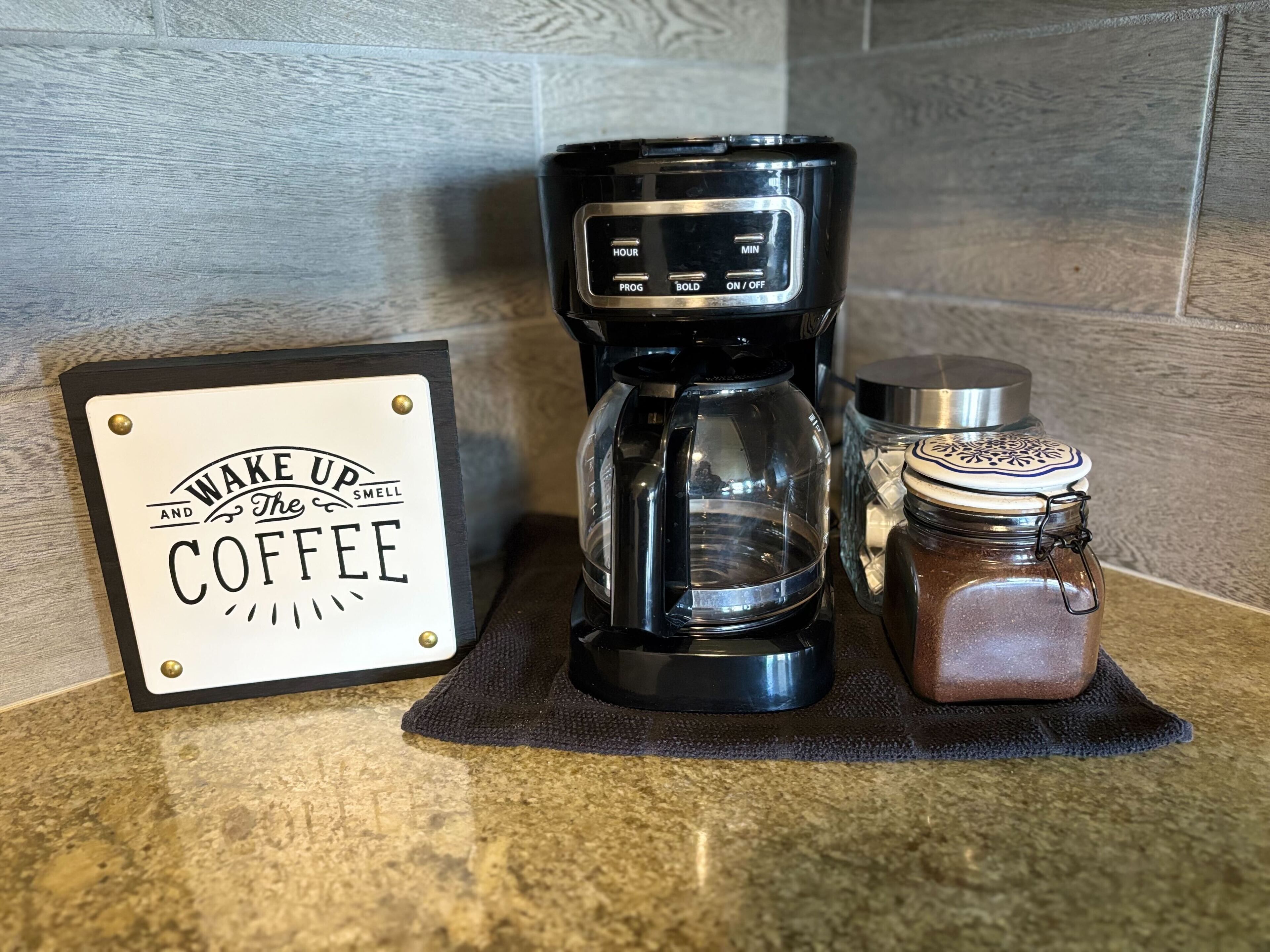 Une machine à café, un bocal de café et un panneau sur lequel on peut lire « Réveillez-vous et profitez de l’arôme du café », le tout posé sur un plan de travail de cuisine.