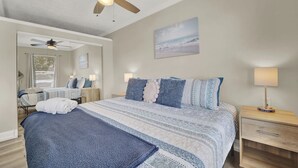 A bedroom with a bed, a ceiling fan, a bedside table with a lamp, a mirror, and a window with blinds.