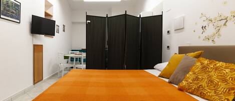A hotel room with a large bed, a TV mounted on the wall, a desk with a chair, and a wardrobe with a mirror.