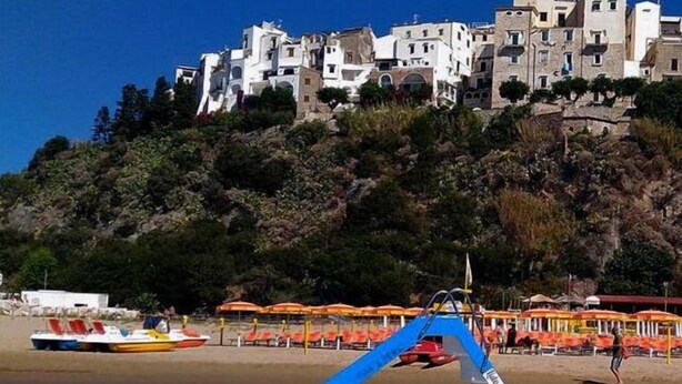 Un barco fondeado cerca de una playa, con edificios blancos y una ladera rocosa al fondo.