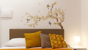 Un dormitorio con cama, almohadas, una mesita de noche, una lámpara y un mural con un árbol y un pájaro.