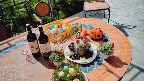 Una mesa dispuesta con un pastel decorado, huevos y botellas de vino, rodeada de sillas y follaje.