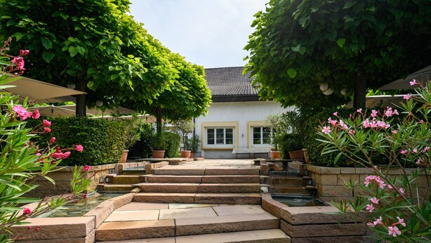 A stone staircase leading to a house with a tiled roof, surrounded by lush greenery and flowering plants.