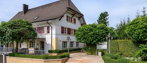 A two-story building with a tiled roof, a balcony, and a well-maintained garden with hedges and trees.