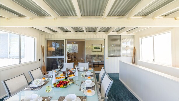 Un comedor con una larga mesa dispuesta para una comida, vista del salón contiguo y un techo con vigas a la vista.