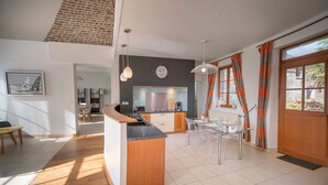 A modern kitchen with a central island, a dining area with transparent chairs, and a view of the outdoors through large windows. - Villa du Ridin - La Villa 4 Fleurs (Le Crotoy)