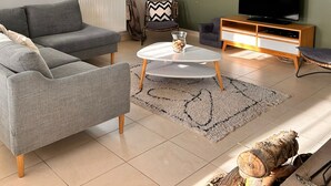 A modern living room with a grey sofa, a round coffee table, a television on a wooden stand, and a large window with curtains. - Villa du Ridin - La Villa 4 Fleurs (Le Crotoy)
