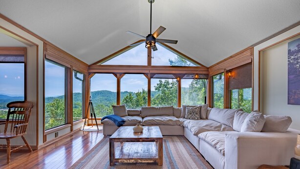 A spacious living room with a large sectional sofa, a wooden coffee table, and a ceiling fan. Large windows offer a view of the outdoors. - Mountain View/Jacuzzi/ Fire pit/ Close to Downtown. Pet Friendly!  (Murphy)