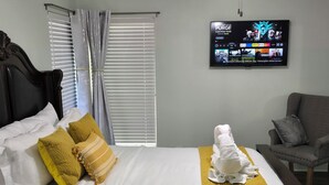 A hotel room with a bed, a grey armchair, a TV displaying a movie, and a window with blinds.