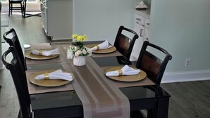 A dining area with a long table set for six, surrounded by chairs, with a kitchen in the background and a large window providing natural light.