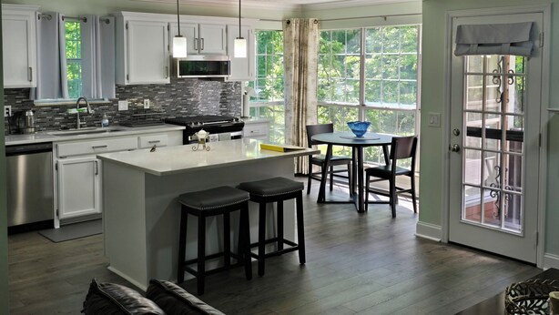 A modern kitchen with a central island, bar stools, and a dining area with a view of greenery.